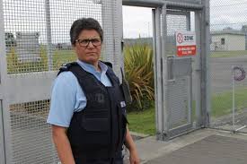 Leonie Aben stands in front of a steel wall surrounding a prison. She is wearing the blue Corrections uniform.